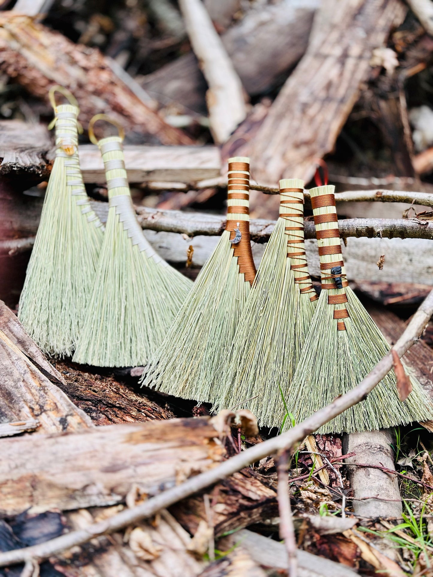 Five hand brooms with crystals and moon pendants attached set amongst a woodpile in the forest